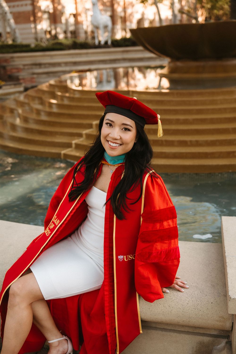 Graduate in red academic regalia and cap sits on stone steps near reflecting pool at sunset.