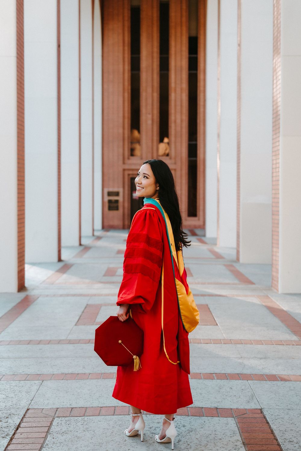 A person in a vibrant red coat poses against a modern architectural backdrop with columns and arches.