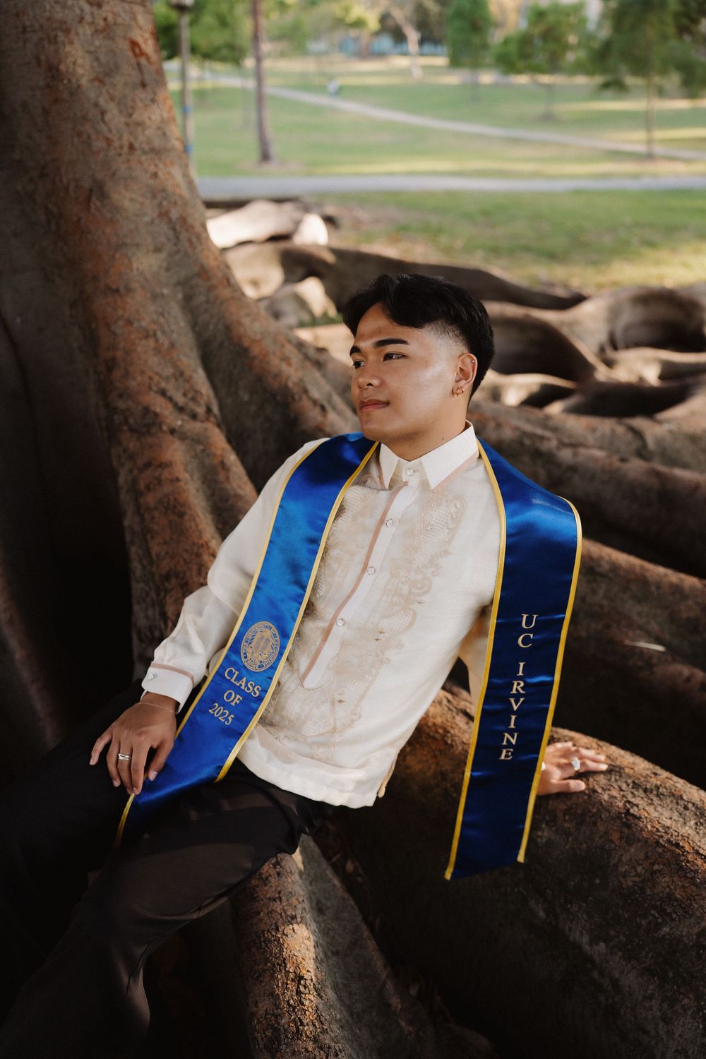 Graduate in white traditional attire and blue sash poses on tree roots during outdoor photo session.