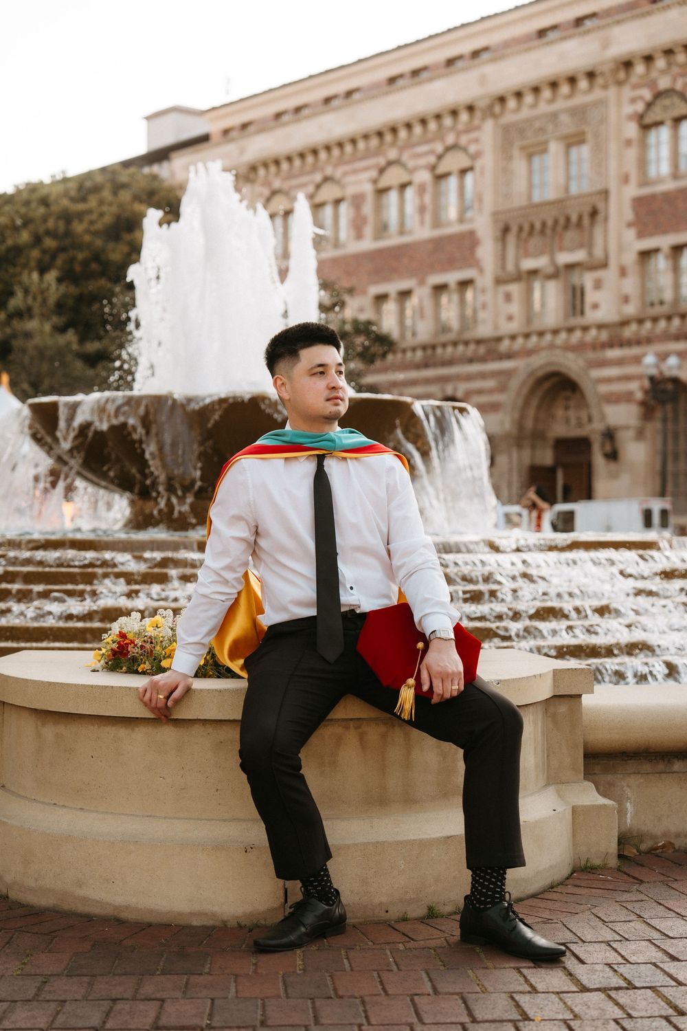 A graduate in formal attire sits on a stone fountain ledge in front of a grand university building.