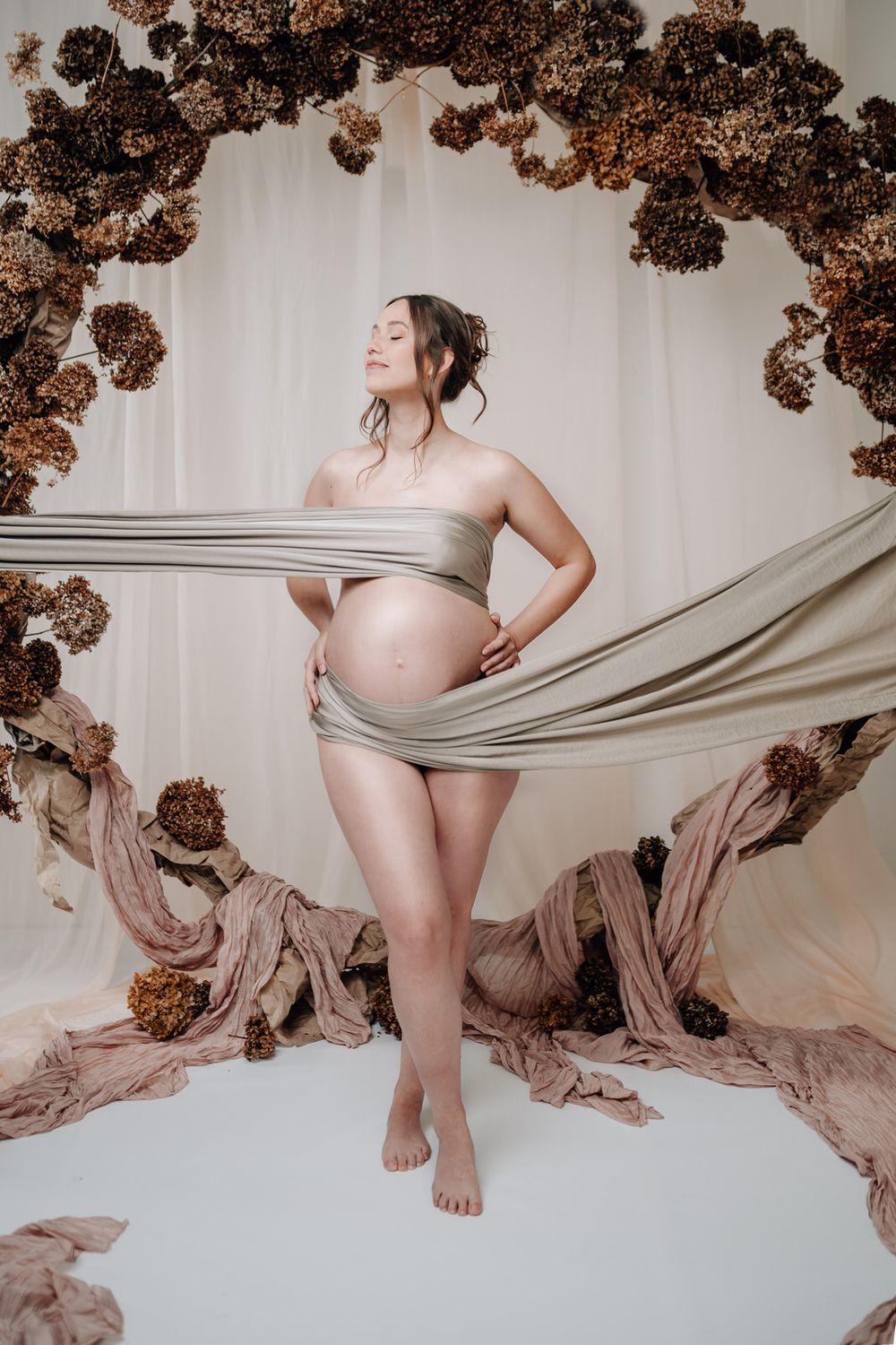 Artistic maternity photo with draped fabric and dried brown floral arch against white backdrop.Babybauch  Fotografie Hessen