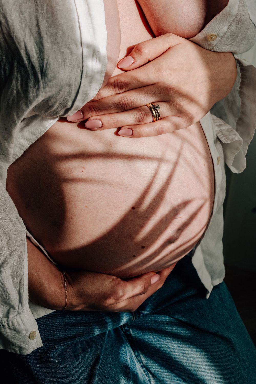 Close-up of pregnant belly with hands gently resting on top, wearing a white shirt and blue pants in soft lighting.Babybauch  Fotografie