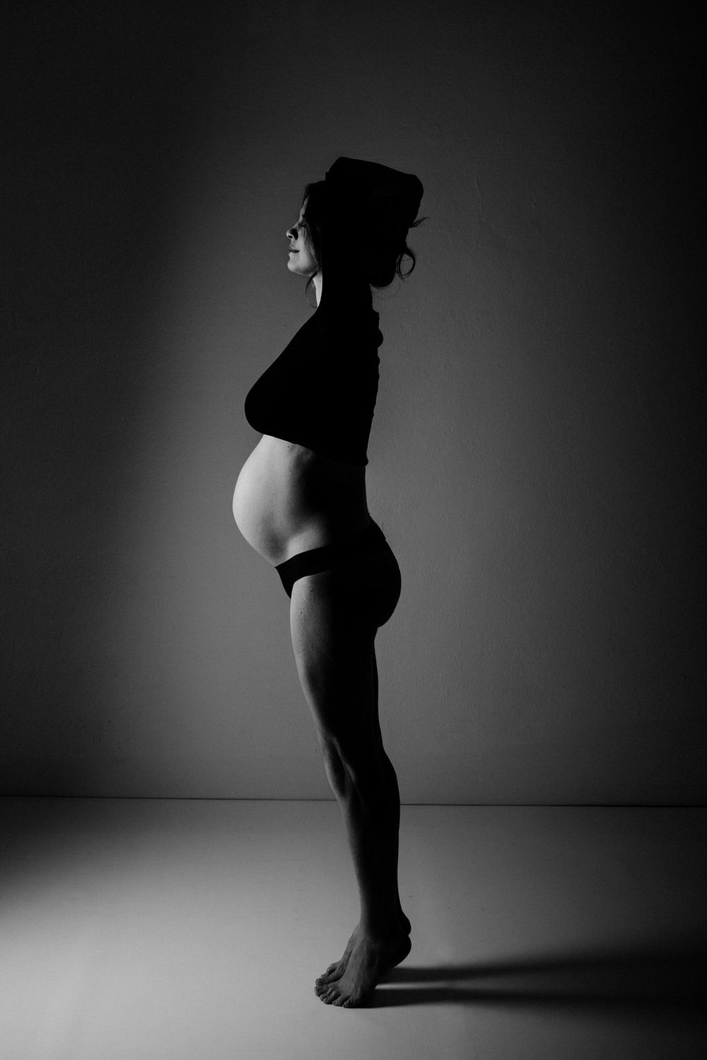 Black and white silhouette of a pregnant figure standing in profile against a dark background.Babybauch  Fotografie Hessen
