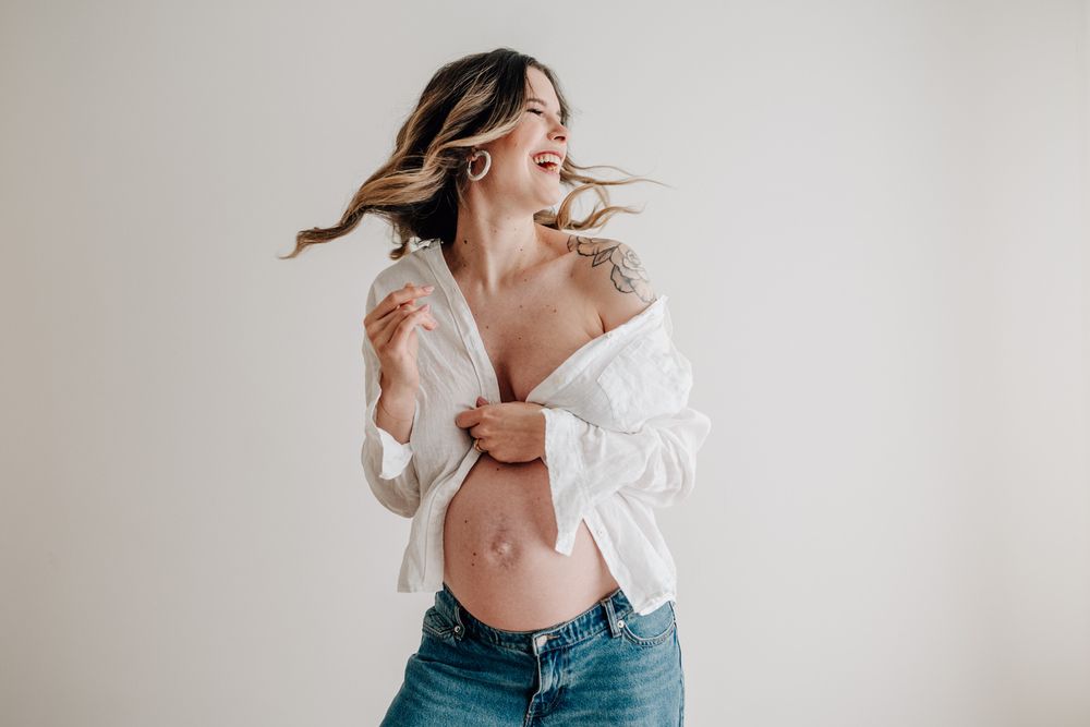 An expectant mother in white shirt and jeans poses playfully against a neutral background.Babybauch  Fotografie Hessen
