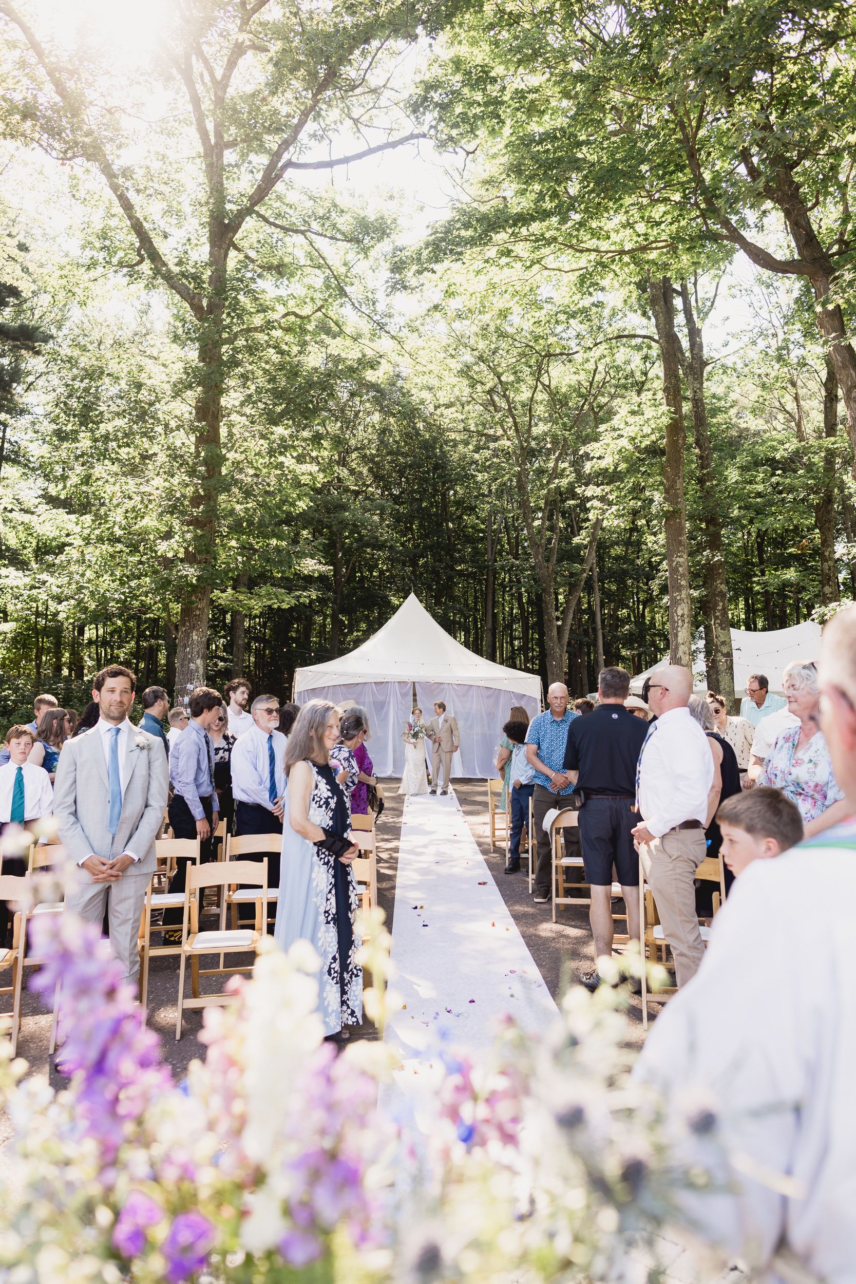 A romantic outdoor wedding ceremony in a wooded setting with a white tent altar and purple wildflowers lining the aisle.