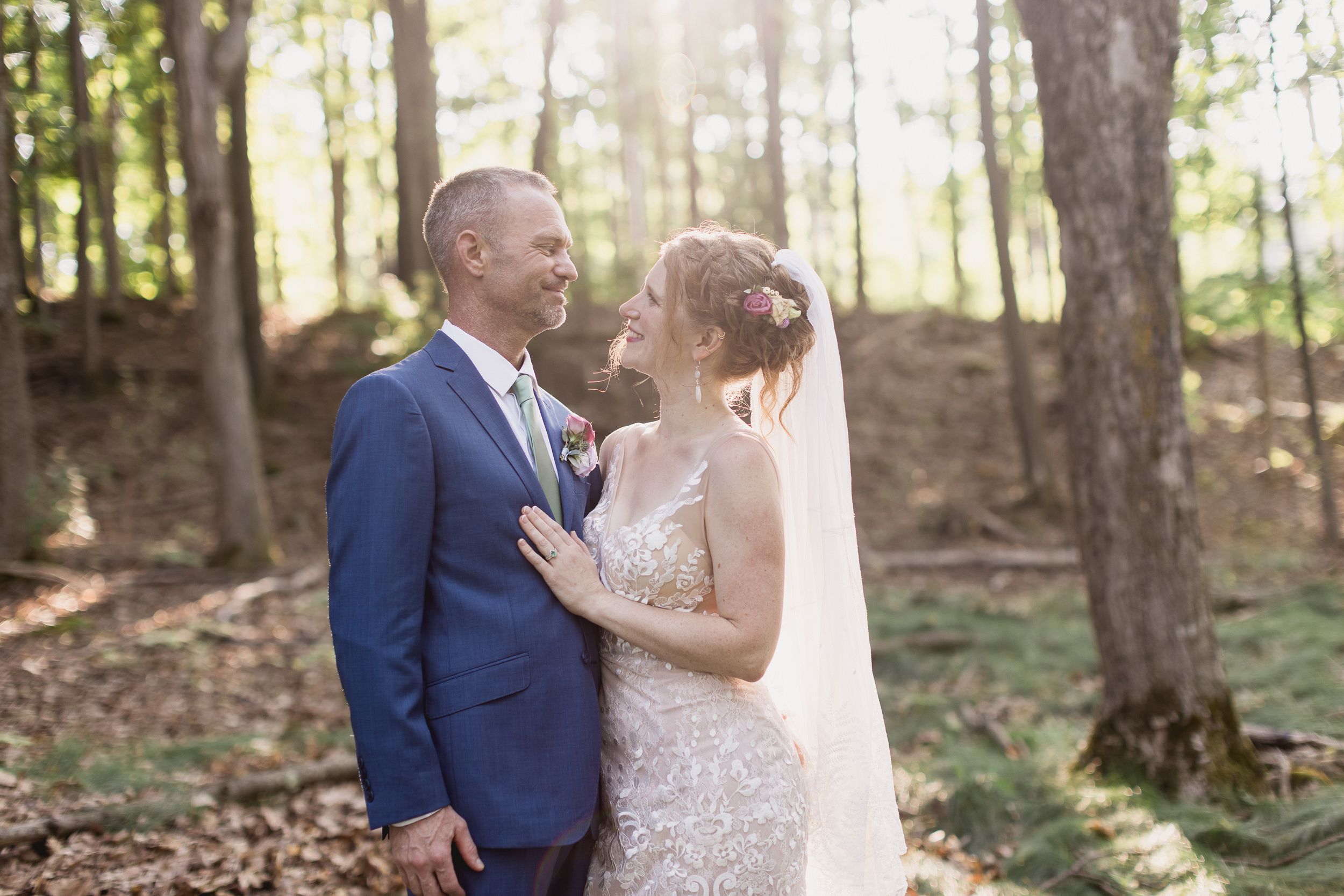A romantic wedding couple shares an intimate moment in a sunlit wooded area during their outdoor ceremony.