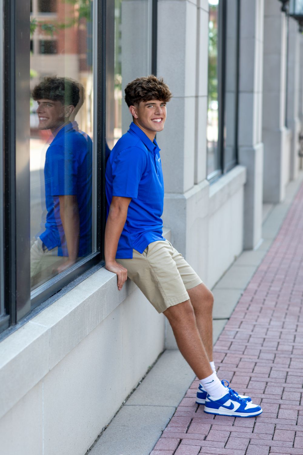 Urban fashion shoot featuring blue Nike polo and white sneakers against reflective windows.