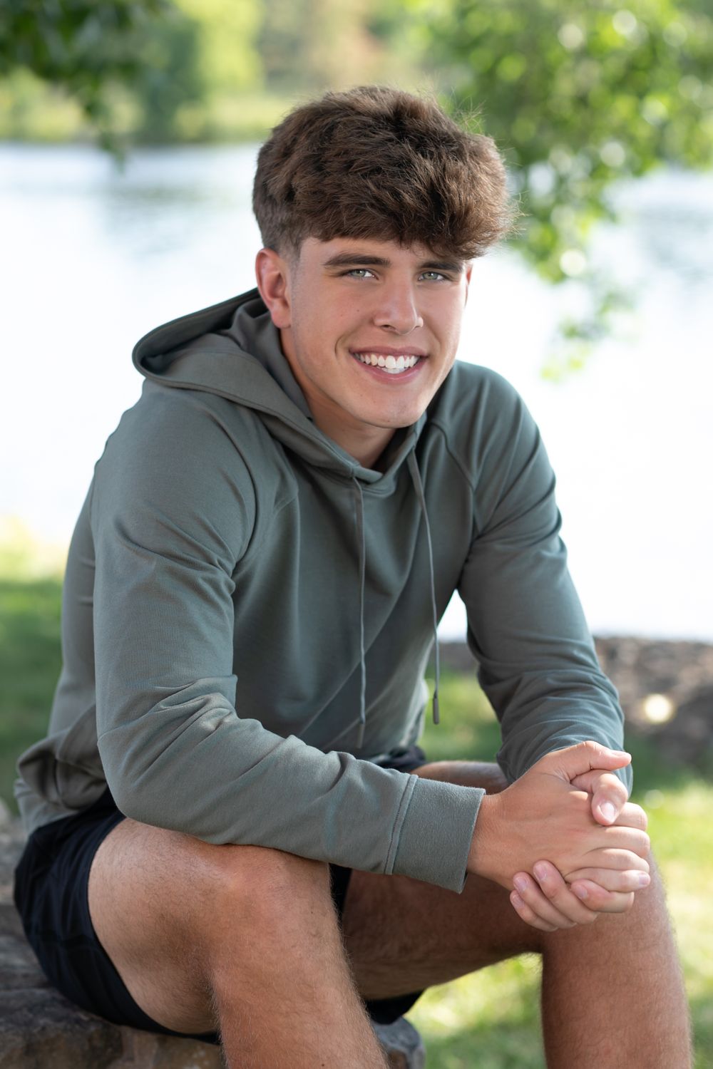 Person in olive green hoodie poses outdoors near a lake on sunny day.