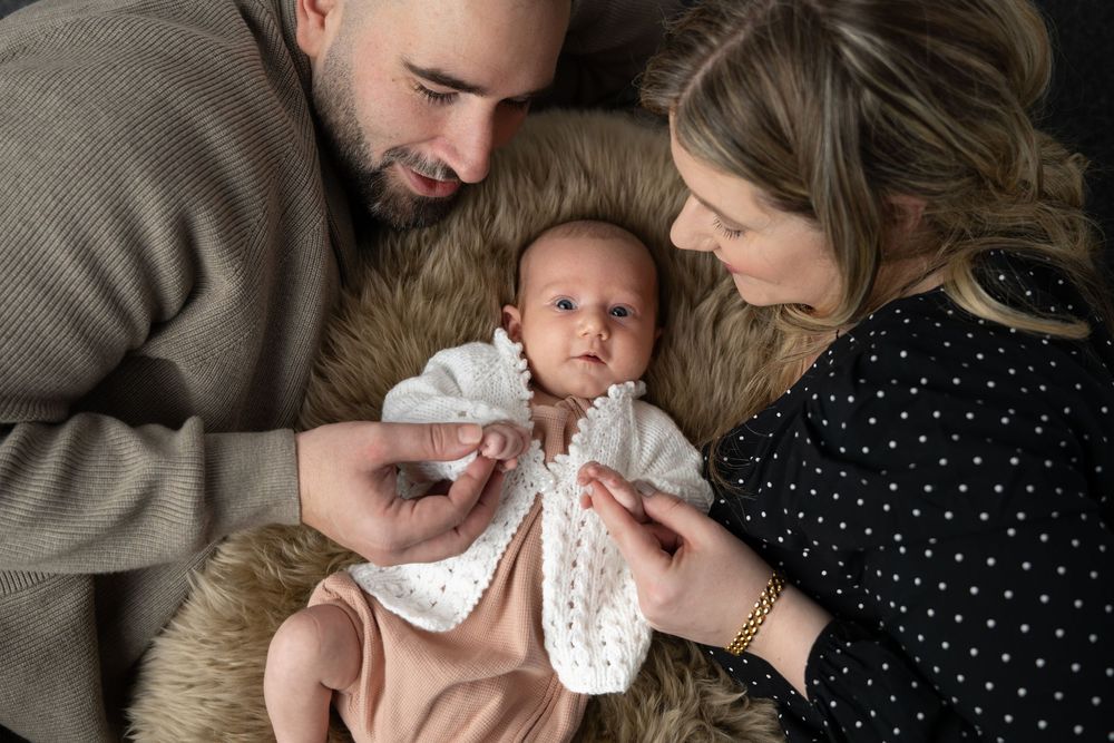 A baby in white clothing lies on a furry blanket while being lovingly attended to by parents.