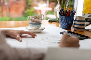 Close-up of hands drawing or writing with teacup and art supplies on desk.