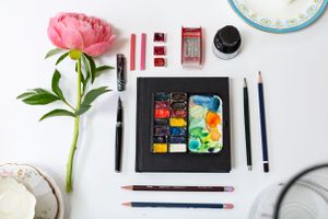 Flat lay of watercolor art supplies with pink peony flower and painting tools on white surface.