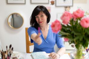 Artist working at desk with pink peonies and wall art in background.