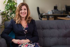 Professional portrait of woman seated on grey couch in office setting.