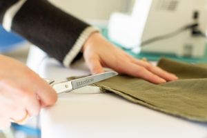 Close-up of hands cutting olive green fabric with scissors on a workspace.