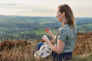 Woman sewing while sitting on grassy hillside overlooking rural valley landscape.