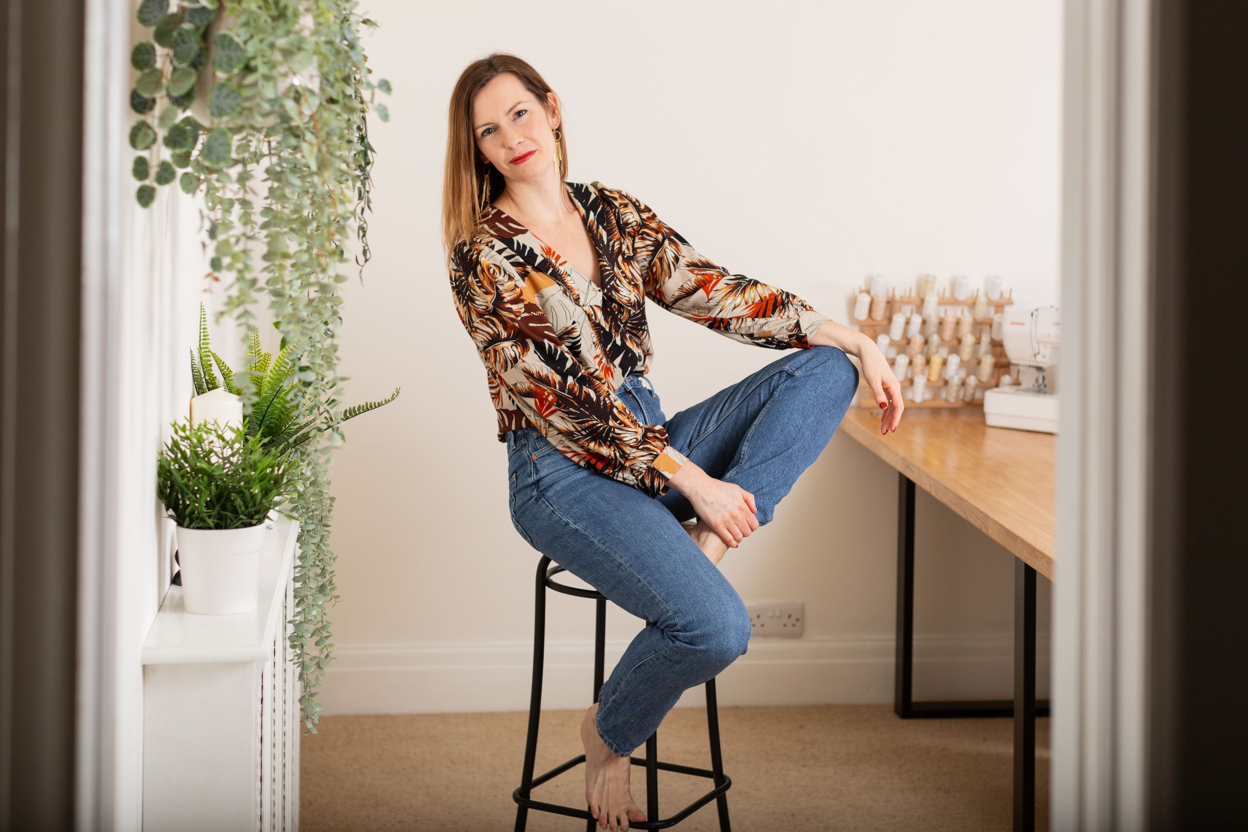 Woman in floral print shirt and jeans poses on barstool in bright interior setting.