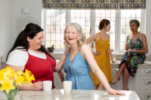 Four women gather around a kitchen counter with yellow flowers and coffee cups.