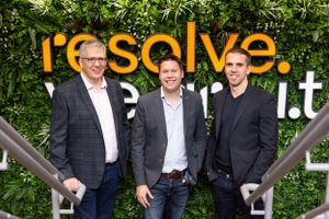 Three professionals standing on stairs in front of a company logo wall with greenery.