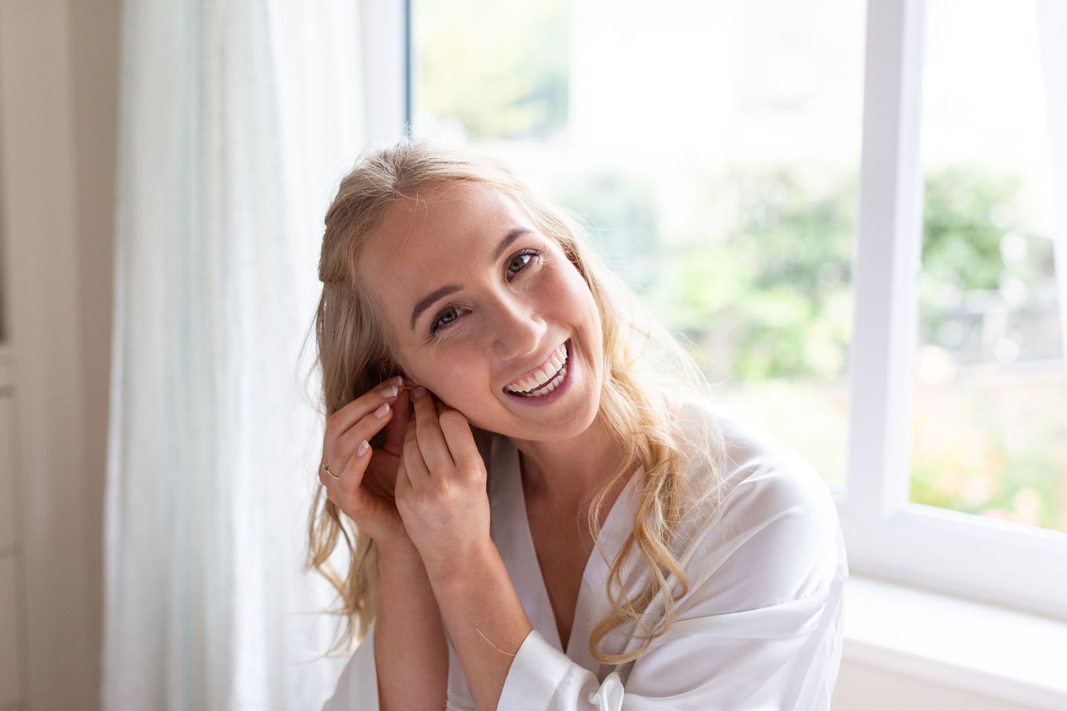 Woman in white robe laughs joyfully near bright window.