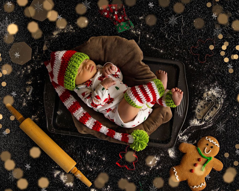 Baby in Christmas pajamas sleeping on baking sheet with gingerbread cookies, rolling pin, and holiday bokeh lights background.