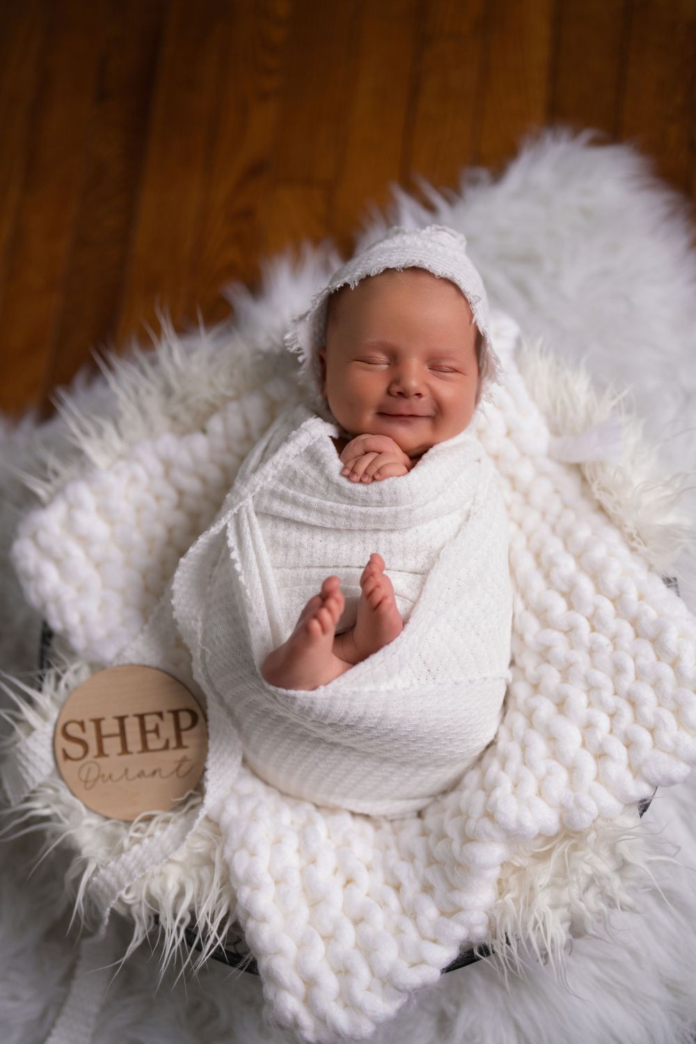 A sleeping newborn swaddled in white on a plush ivory blanket with a wooden round 'SHEP' sign decoration.