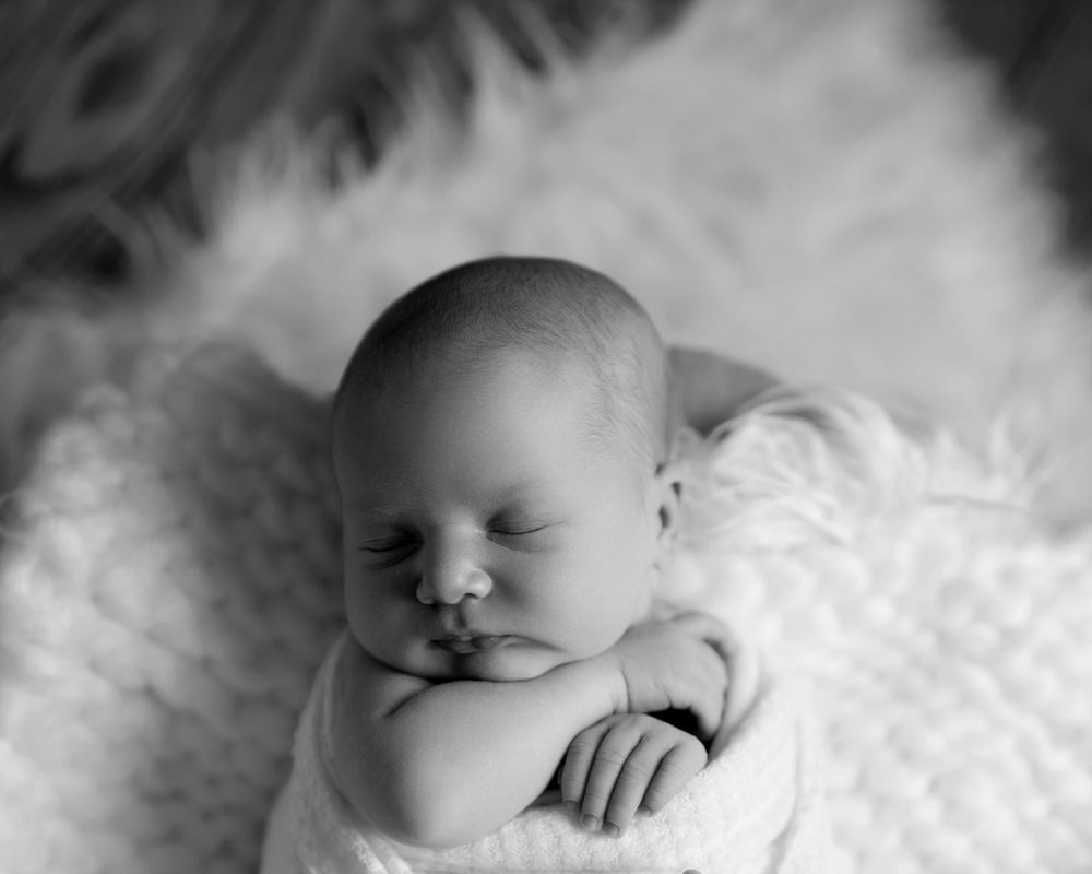 Black and white portrait of a sleeping newborn baby resting on soft white textured blankets.