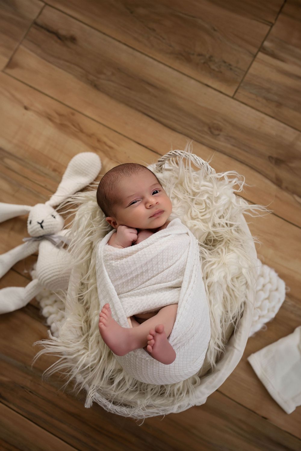 Newborn baby wrapped in white swaddle blanket lying on fluffy white rug with plush bunny toy on wooden floor.