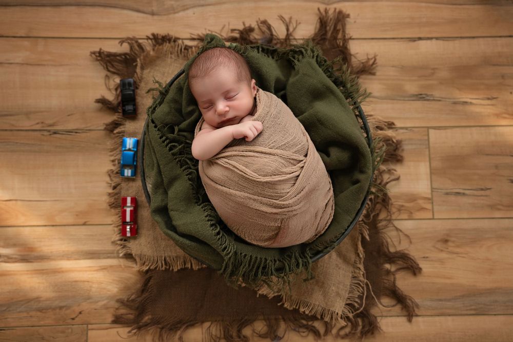 A swaddled newborn sleeps peacefully on a green pillow surrounded by rustic textures on wooden floor.