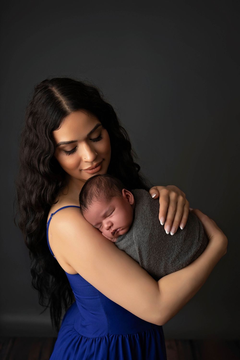 A tender moment captured in a portrait as a woman in a blue dress embraces a sleeping newborn wrapped in gray.