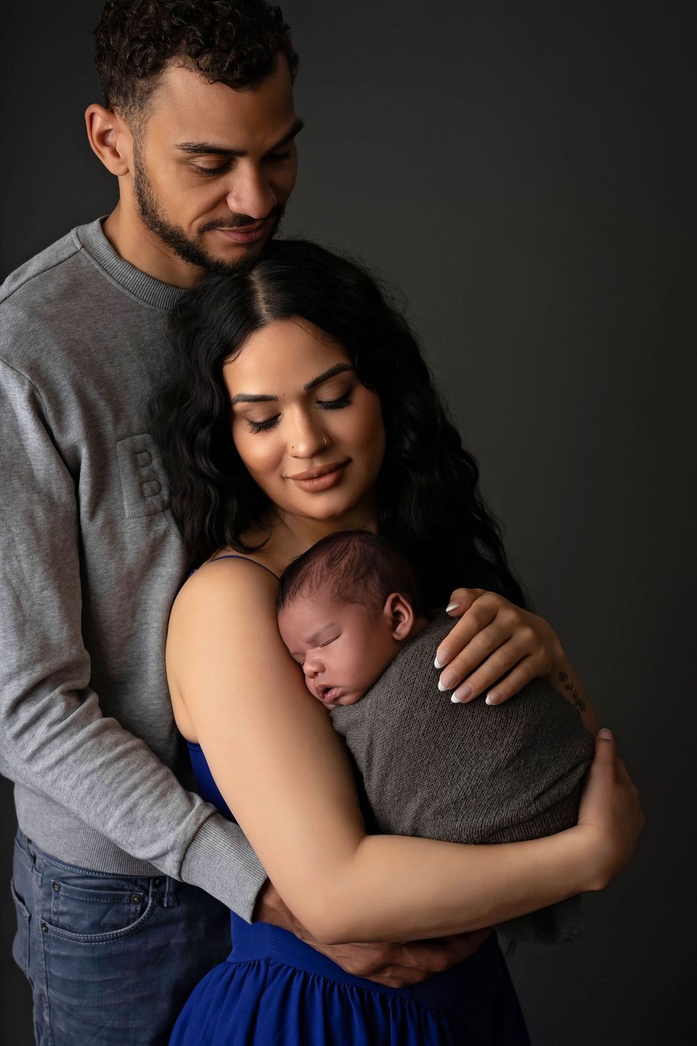A tender family moment as parents embrace their sleeping newborn baby against a dark background.