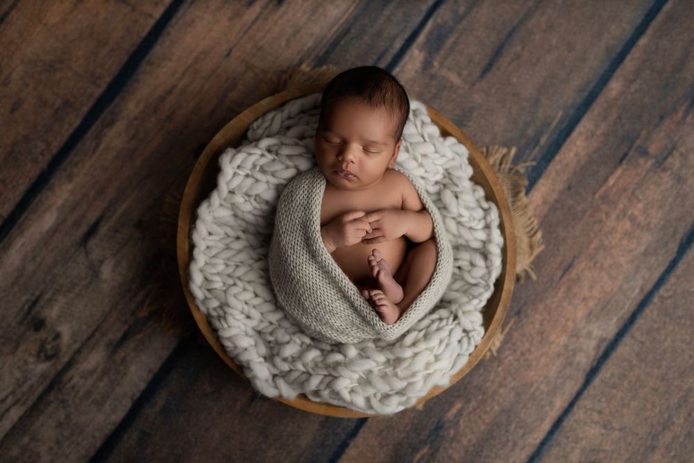 Newborn baby sleeps peacefully in round basket with chunky knit blanket on wooden floor.
