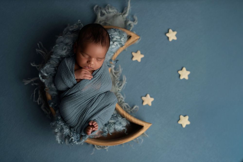 Sleeping newborn wrapped in blue swaddle lying on decorative wooden crescent moon with star decorations against blue backdrop.
