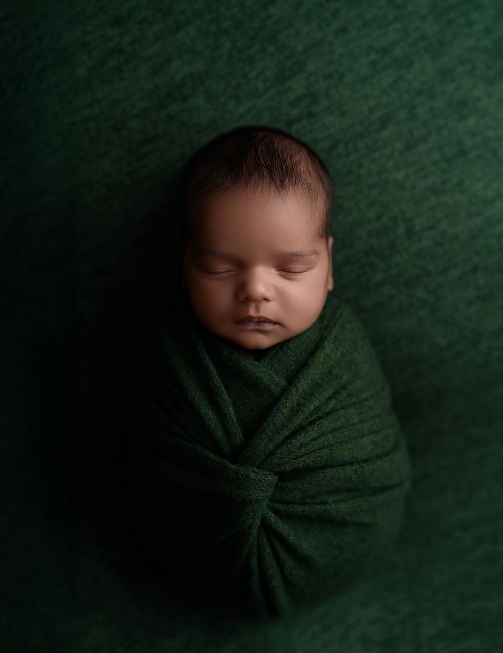 Sleeping newborn baby wrapped in deep green swaddle blanket against matching green textured backdrop.
