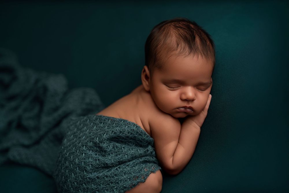 Sleeping newborn baby wrapped in teal knit fabric against dark teal background.