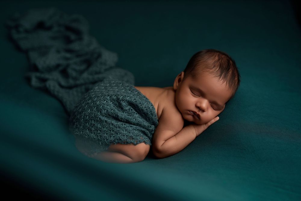 Sleeping newborn wrapped in gray knit blanket on dark teal background in artistic portrait.