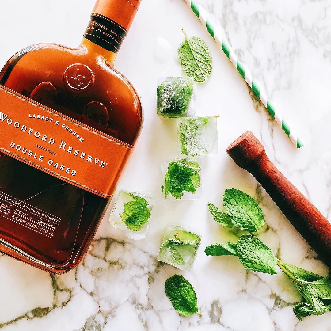 Bourbon bottle with mint leaves, ice cubes, and striped straws on marble surface.
