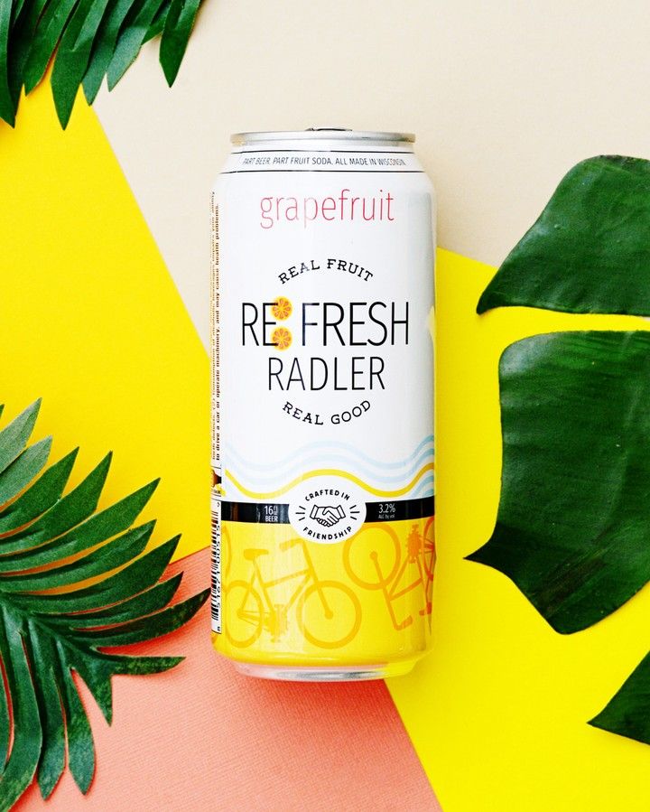 White beverage cans with grapefruit radler displayed against tropical leaves on yellow background.