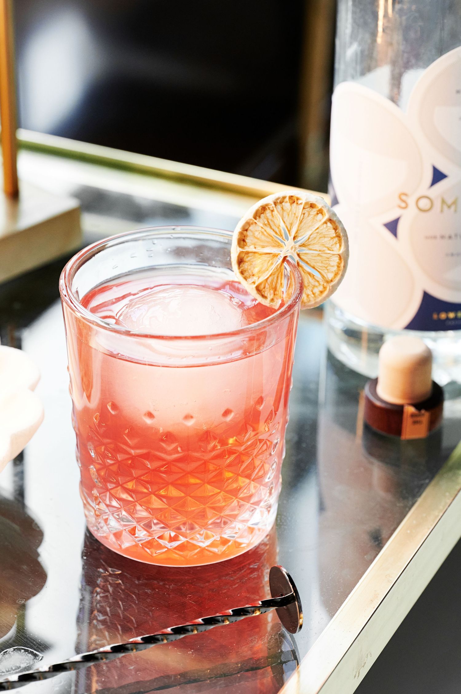 Pink cocktail with dried citrus garnish served in a textured crystal glass with cocktail spoon and bottle.