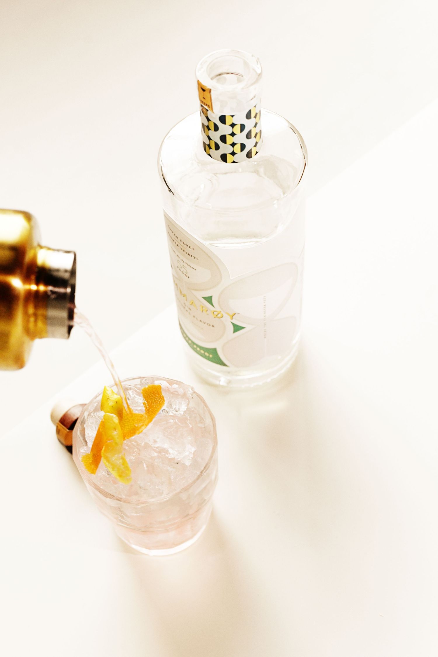 High-key photo of decorative bottle and cocktail with citrus garnish on white surface.