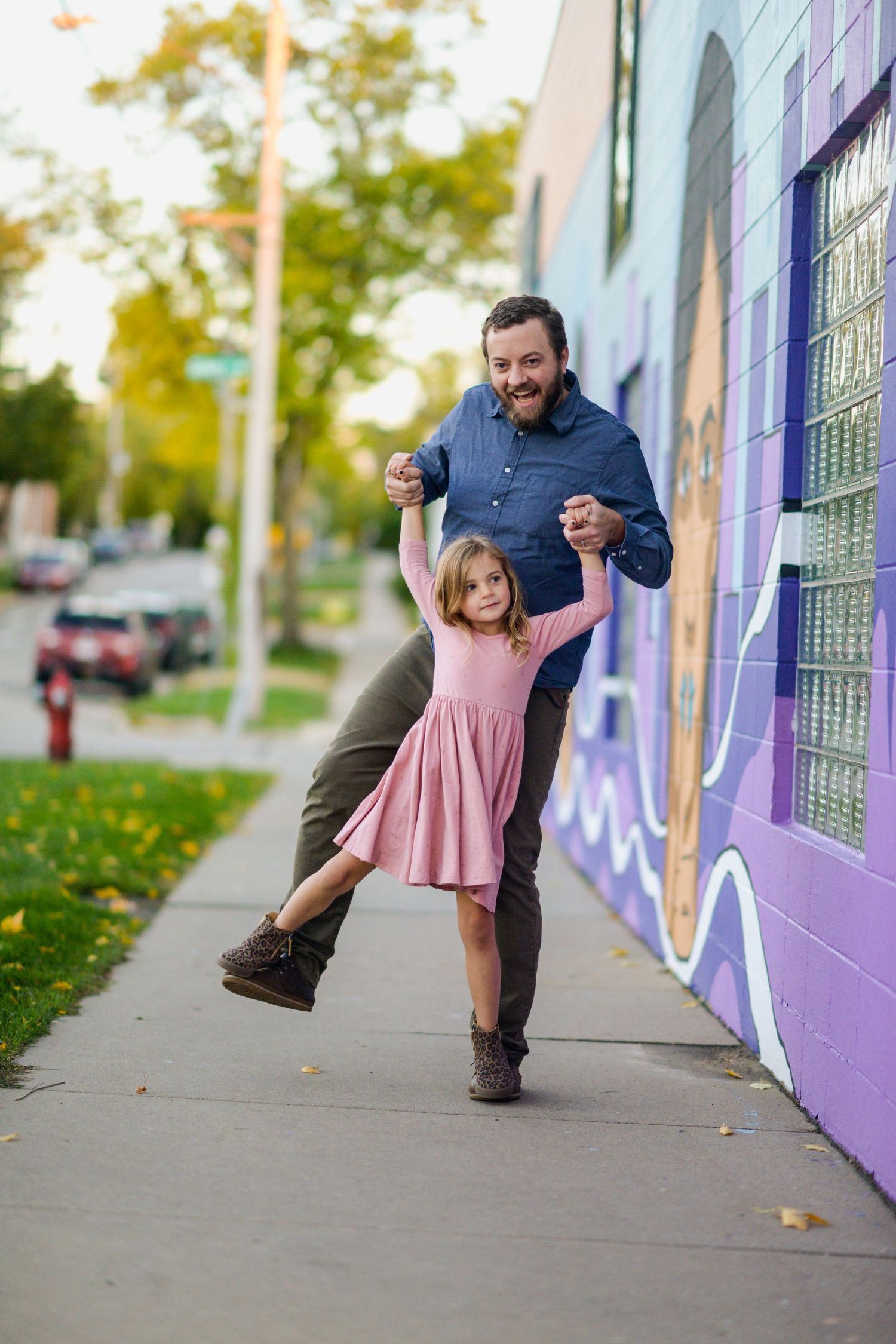 Playful dance pose in front of colorful mural wall with autumn leaves on sidewalk.
