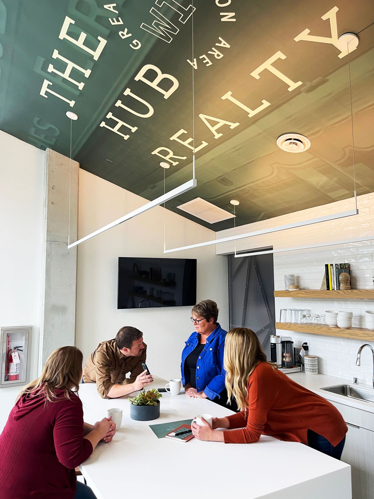 Modern office meeting space with text on ceiling and people gathered around white table.