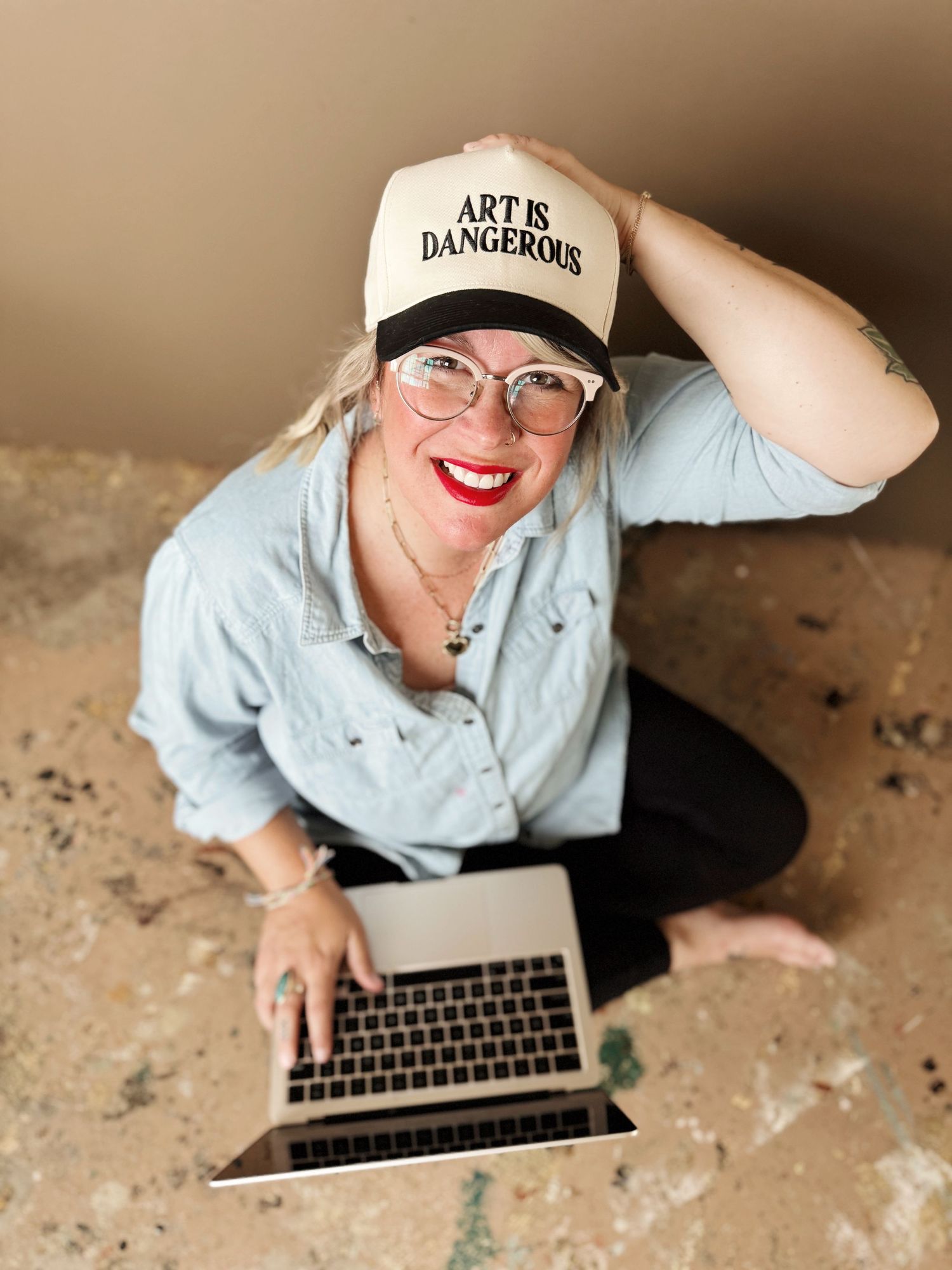 Person wearing 'Art is Dangerous' cap working on laptop in casual setting.