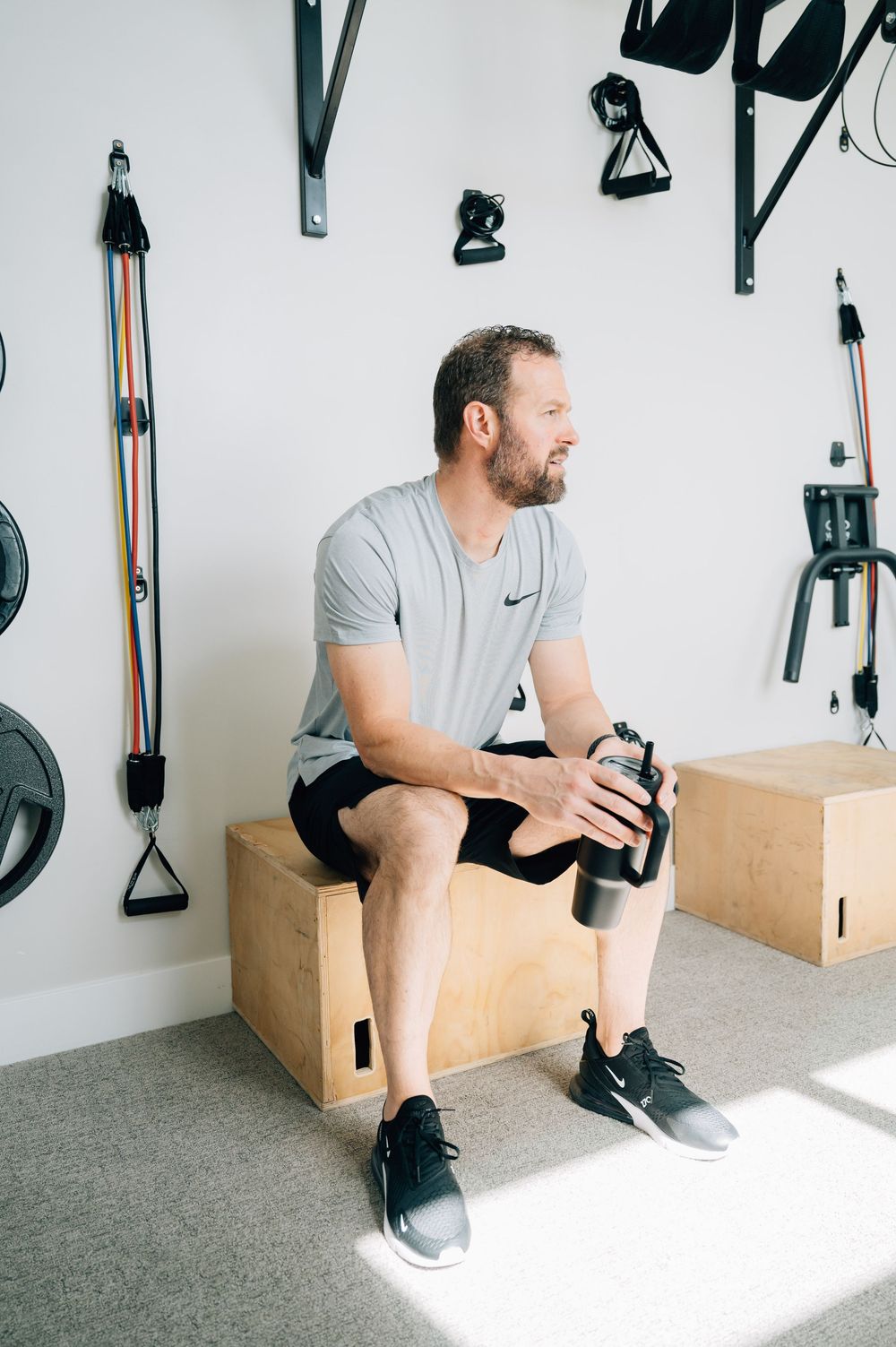 Person in gray Nike shirt sits on wooden box in home gym with resistance bands and workout equipment on wall.