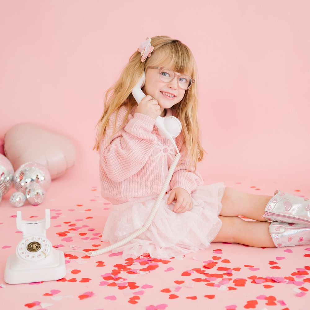 Retro pink telephone scene with scattered red heart confetti and person in cozy sweater posing with vintage phone.