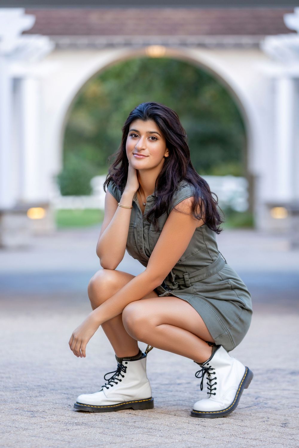 A person in a casual olive dress crouches while wearing white combat boots under an archway.