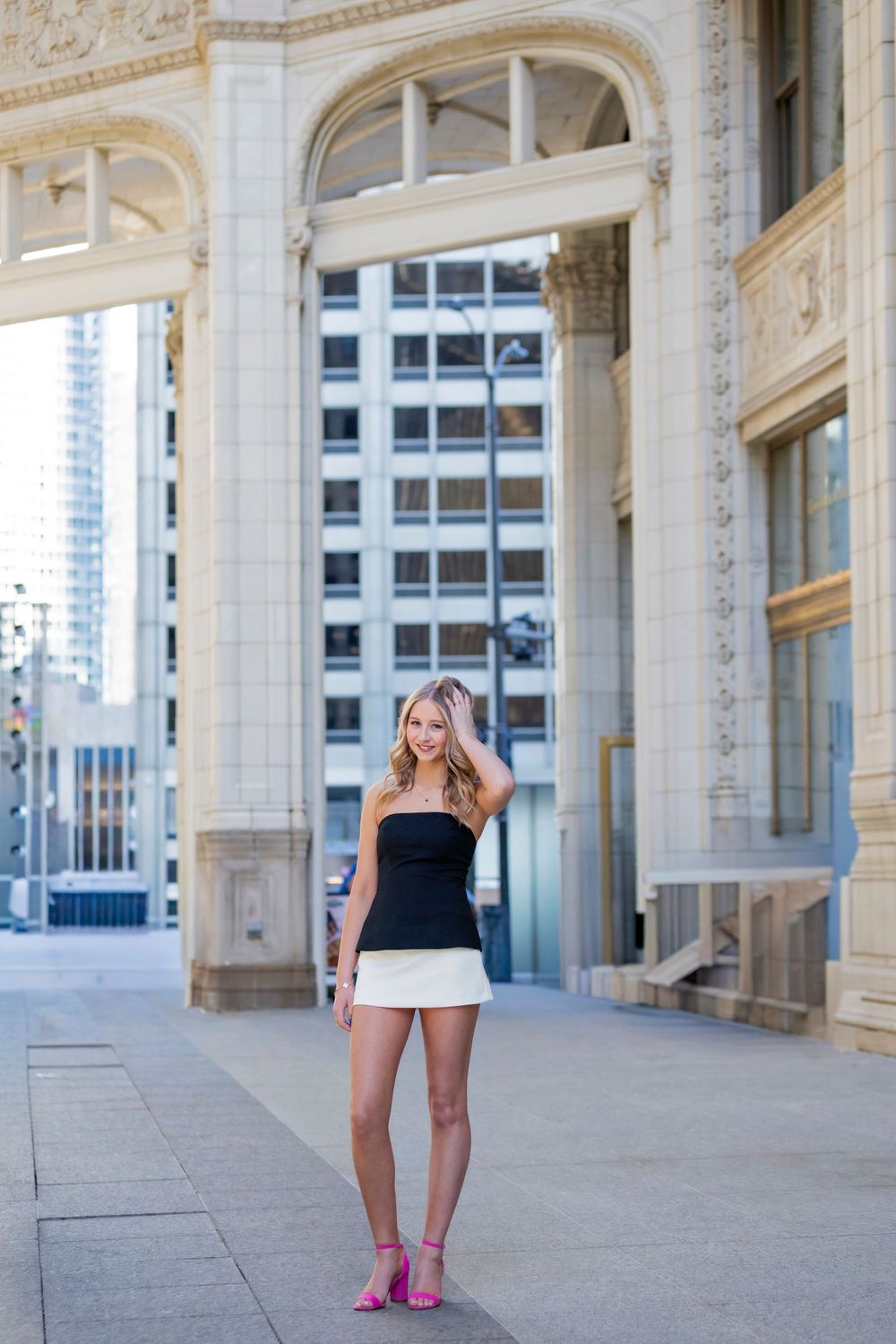 Blonde woman in black tube top and white mini skirt with pink heels posing in front of ornate building.