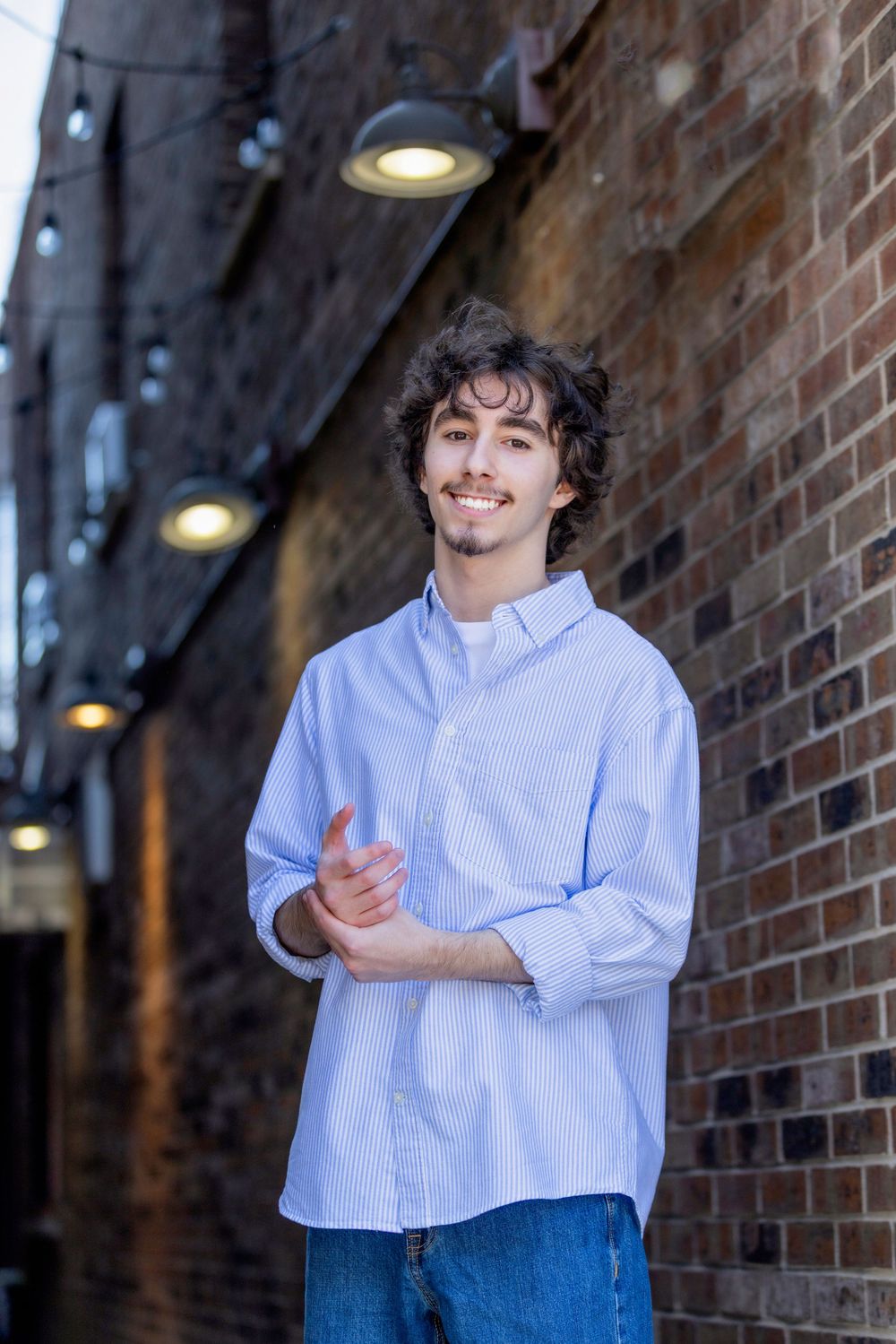 Young man with curly hair smiling in blue shirt and jeans, posing in a brick alley with string lights.
