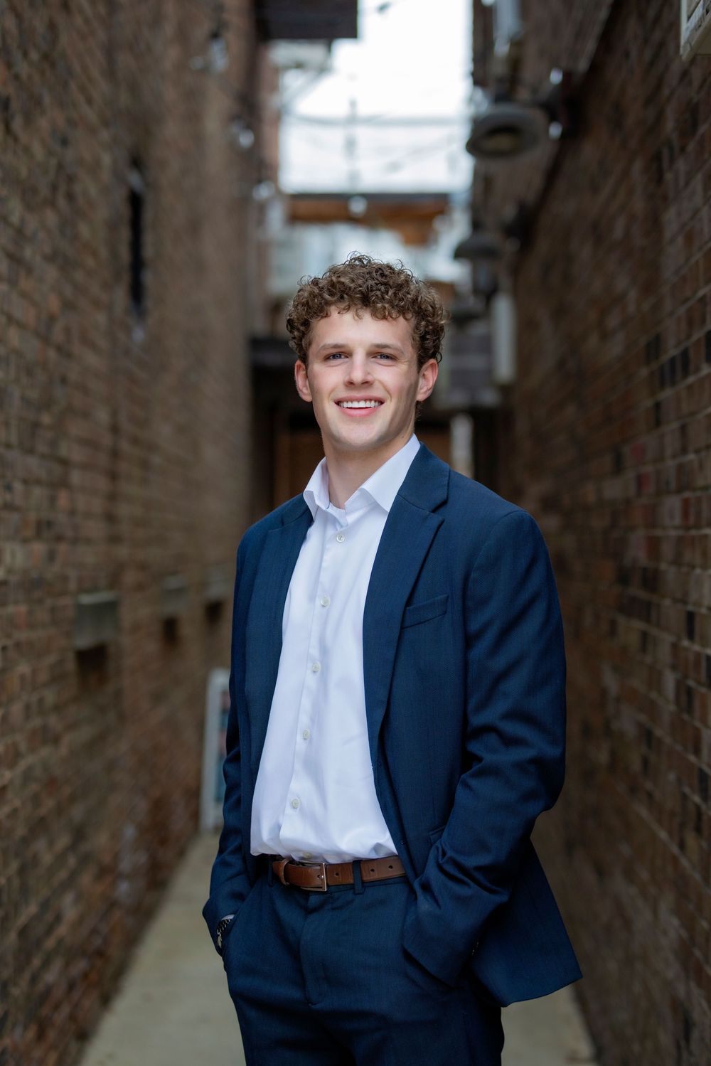 Young man in navy blazer and white shirt smiling in a brick alleyway, ideal for senior portraits.