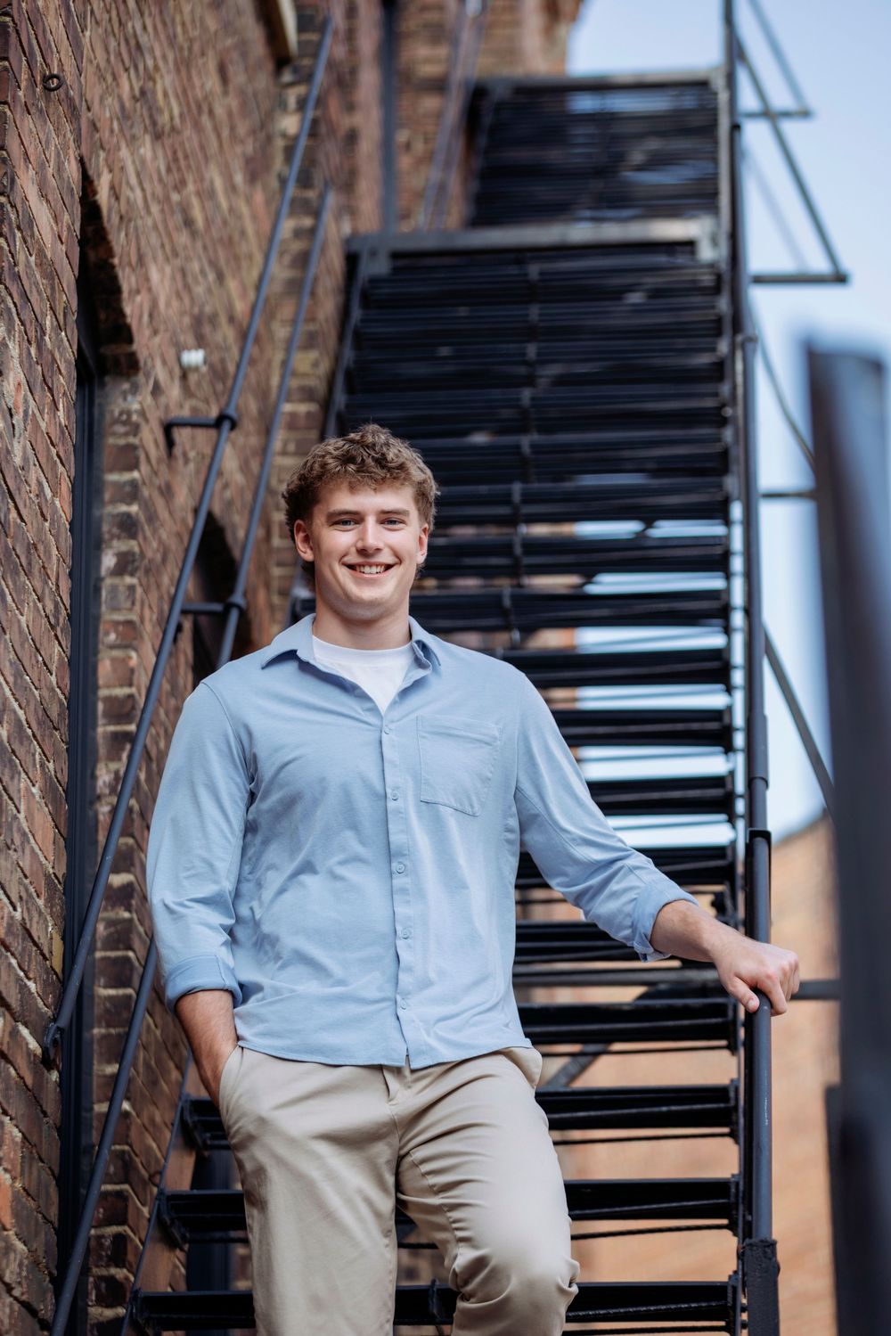 A casual portrait against a brick wall and metal staircase wearing a light blue button-down shirt and khaki pants.
