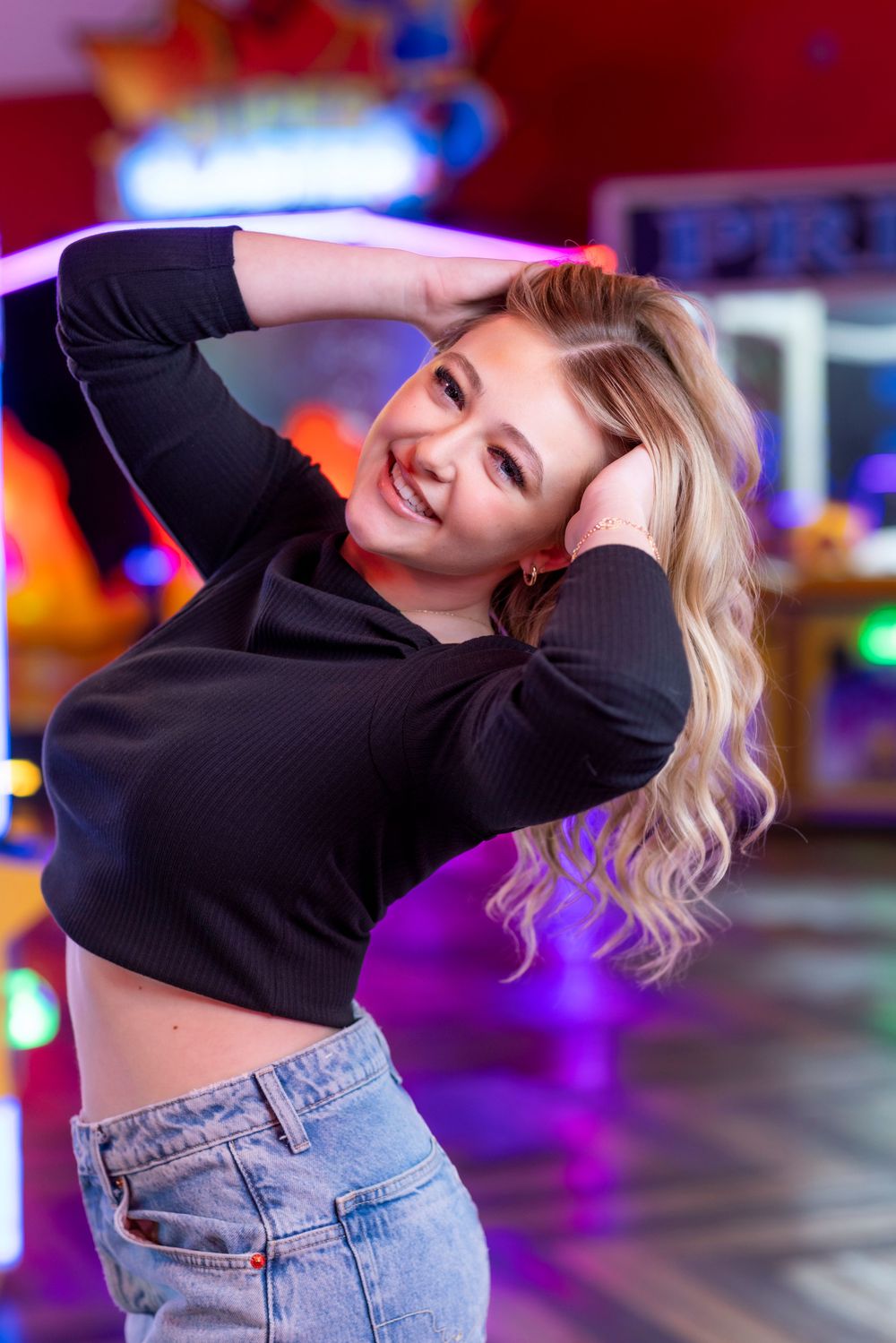 Smiling blonde woman in black crop top and jeans posing in colorful arcade with neon lights.